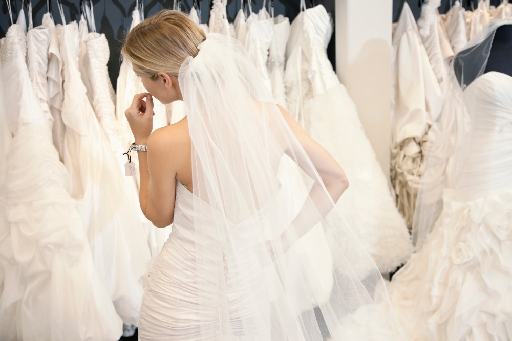 Back view of a young woman in wedding dress looking at bridal gowns on display in boutique